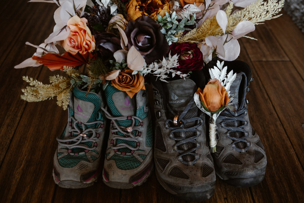 a pair of vasque hiking boots with wedding rings tied into the shoe laces and a floral bouquet behind the shoes