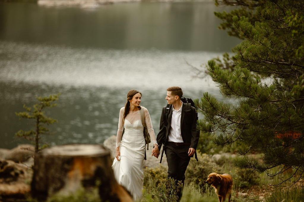 a couple wearing wedding clothes are hiking on a trail holding hands and looking at each other with a lake in the background