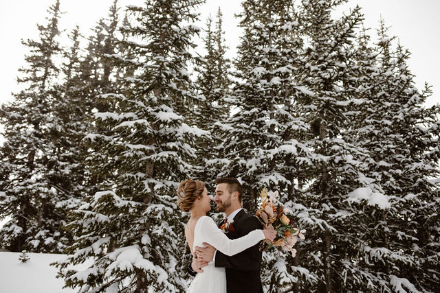 a couple wearing wedding attire is standing in snow wearing snowshoes surrounded by snow covered evergreen trees