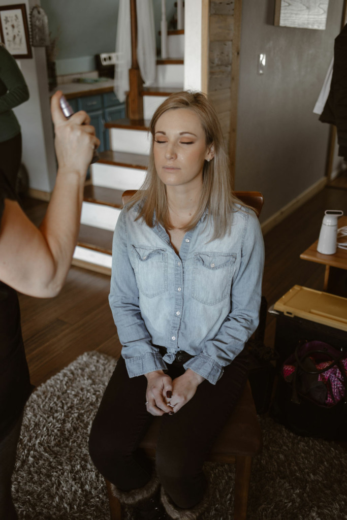 a bride wearing a blue button up is getting her make up professionally done on the morning of her wedding day in the living room of an AriBnb