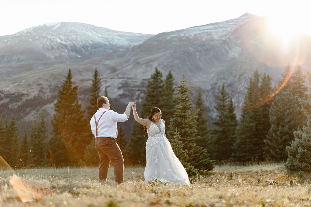 a bride and groom wearing wedding attire is slow dancing in an evergreen forest with a mountain backdrop during sunest