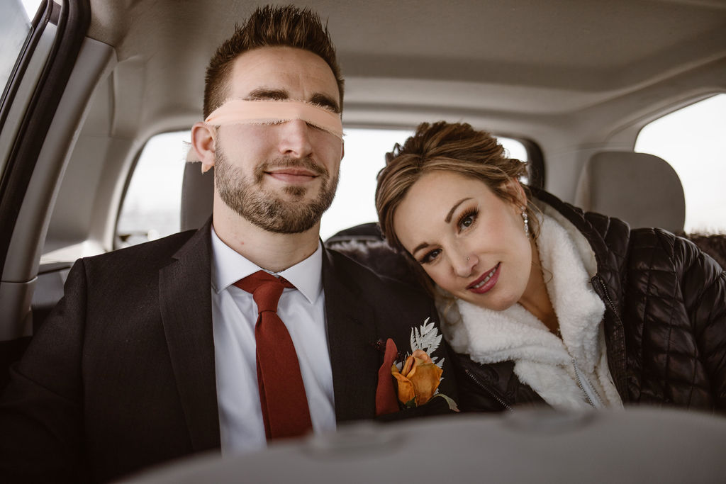 a white male dressed in wedding attire is blind folded sitting in the back of a car while a white female women dressed in wedding attire is sitting next to him smiling