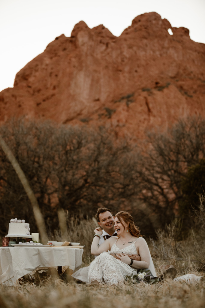 a bride and groom wearing wedding attire are sitting in a grassy area having a picnic at the garden of the gods park