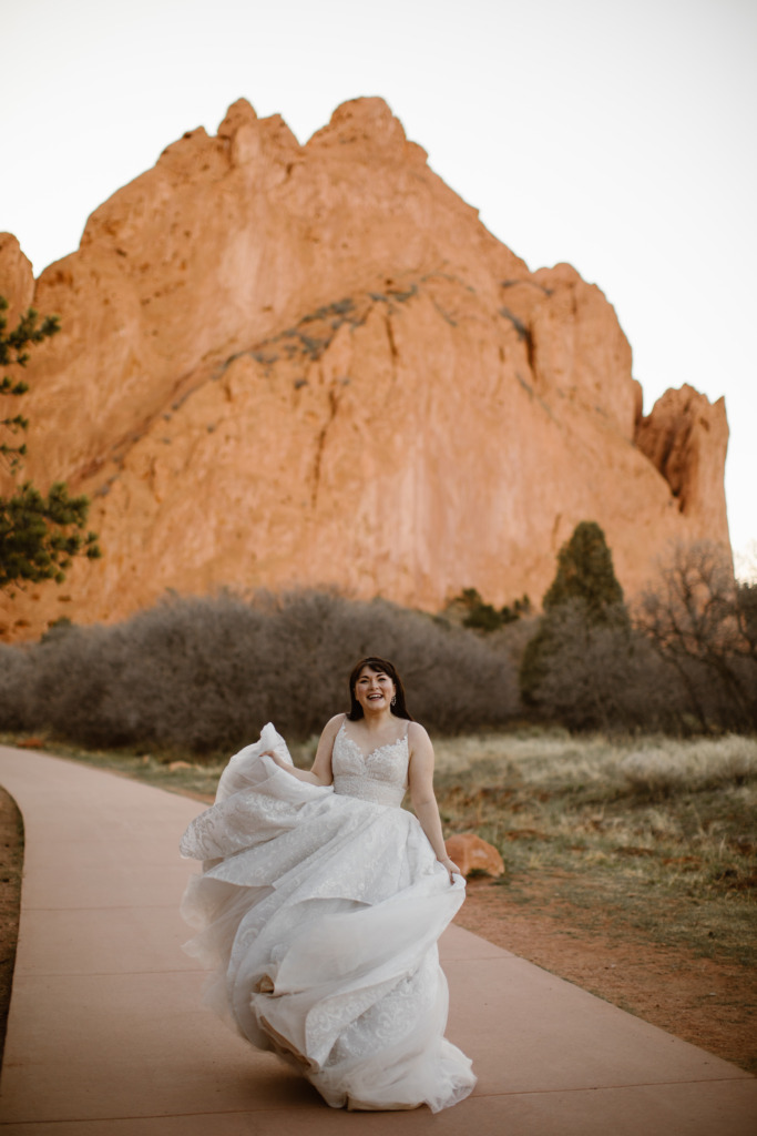 a bride wearing a white wedding gown is dancing on a paved walking path with a big red rock formation in the background at the garden of the gods park
