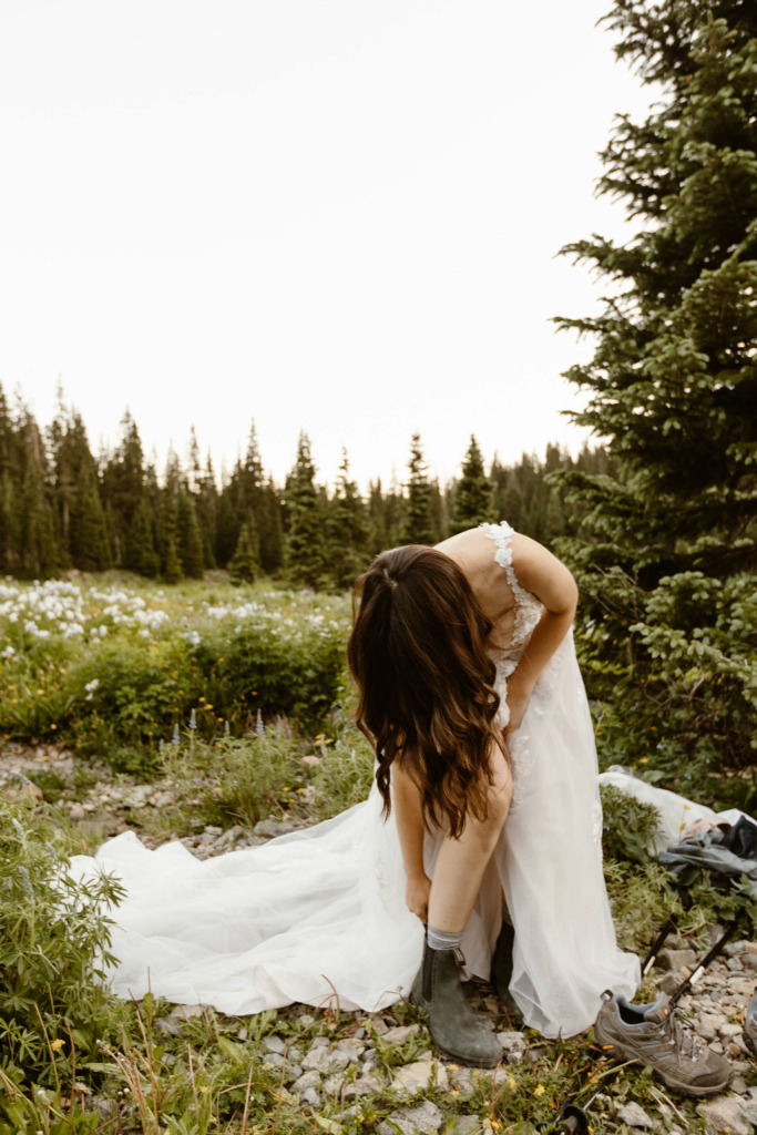 a female bride wearing a white wedding dress is putting on hiking boots in the middle of the woods