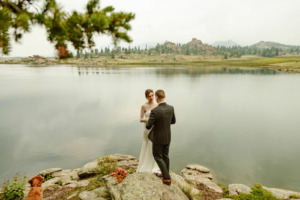 a couple wearing wedding clothes is standing on a rock next to a lake during their elopement ceremony in colorado