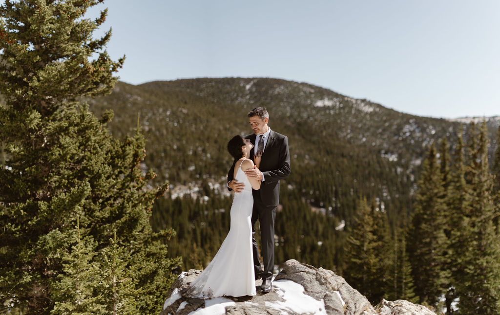 a couple wearing wedding attire are standing on a rock with evergreens in the background during their st marys glacier elopement
