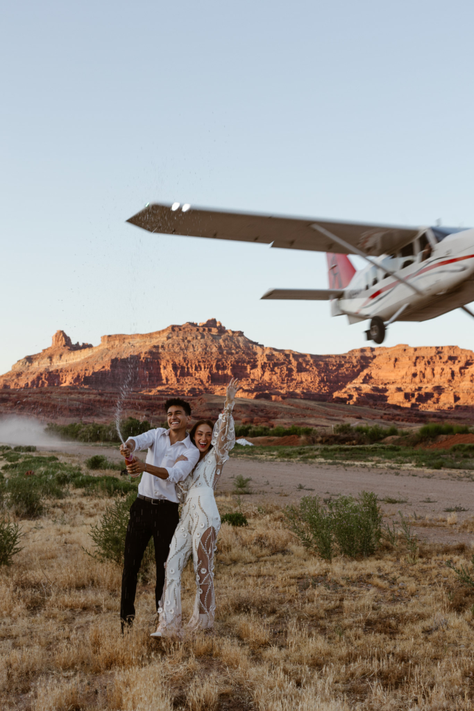 a bride and groom popping a bottle of champagne while a plane flies over their head in the desert