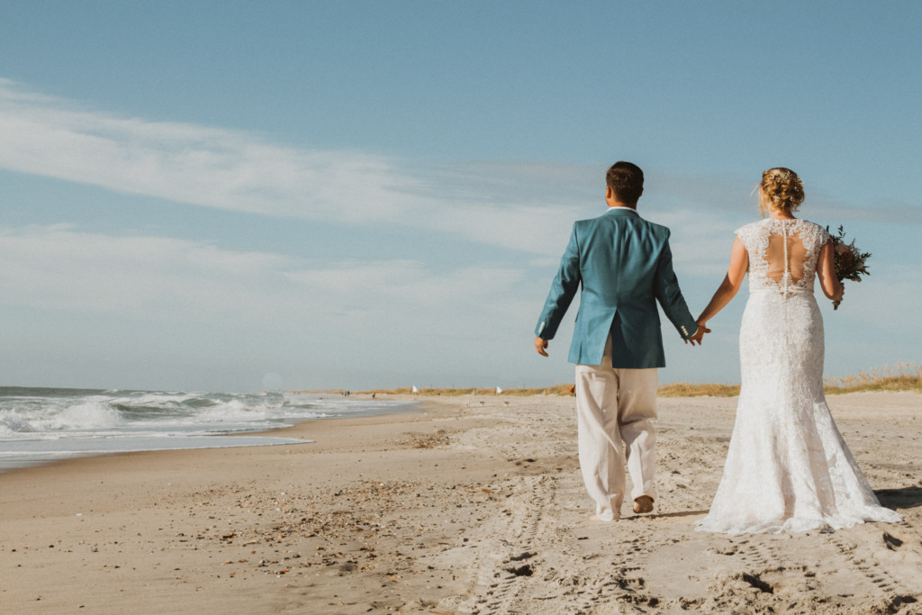 a bride and groom walking hand in hand down the beach away from the camera