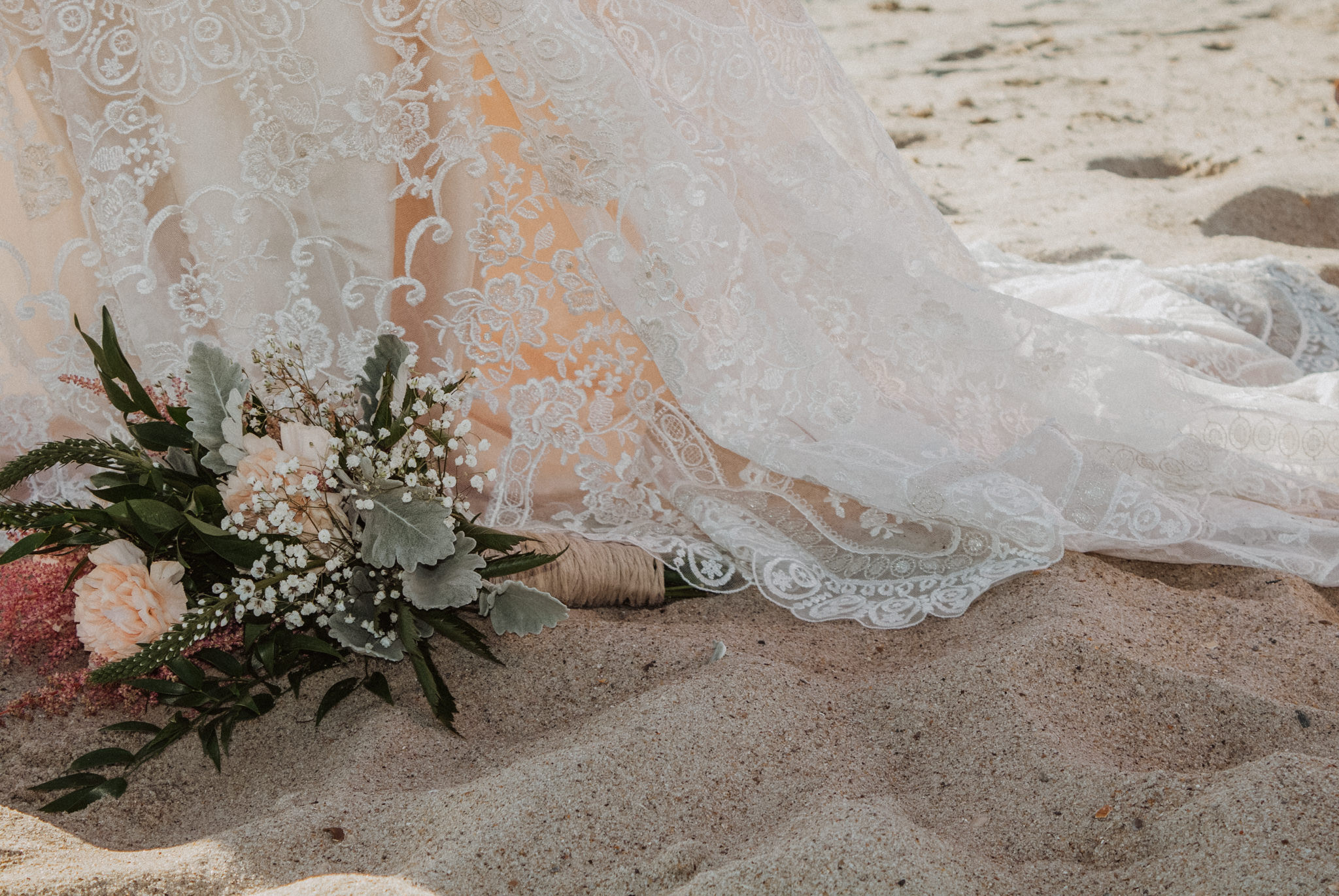 a pink bouquet in the sand next to a lace wedding gown