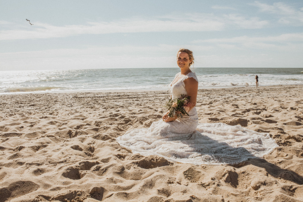 a bride wearing a lace wedding dress sitting in the sand