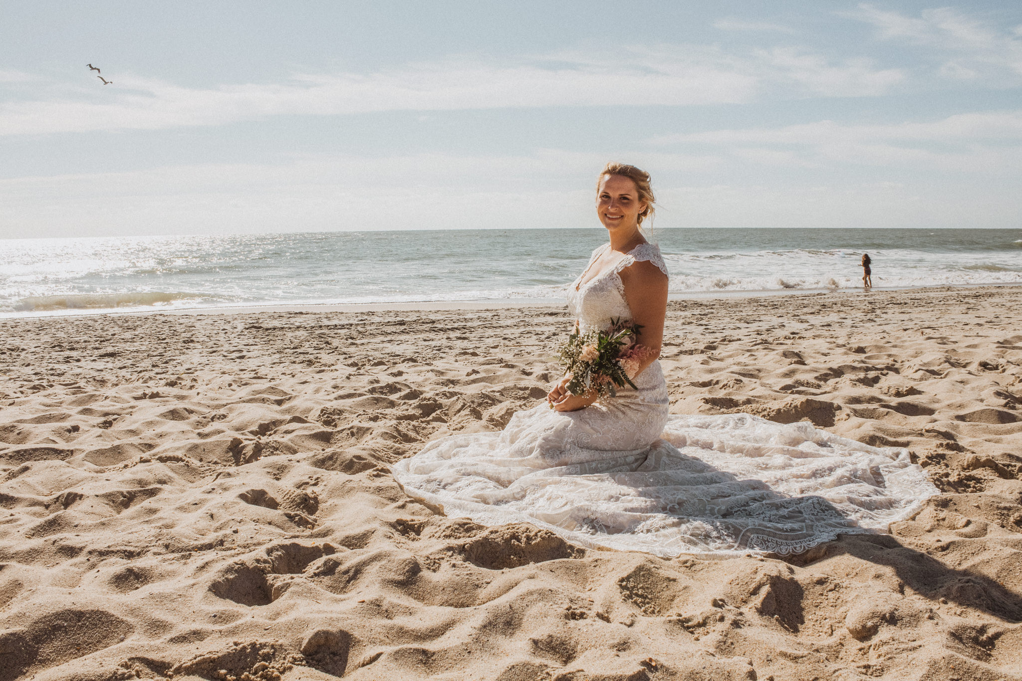 a bride wearing a lace wedding dress sitting in the sand