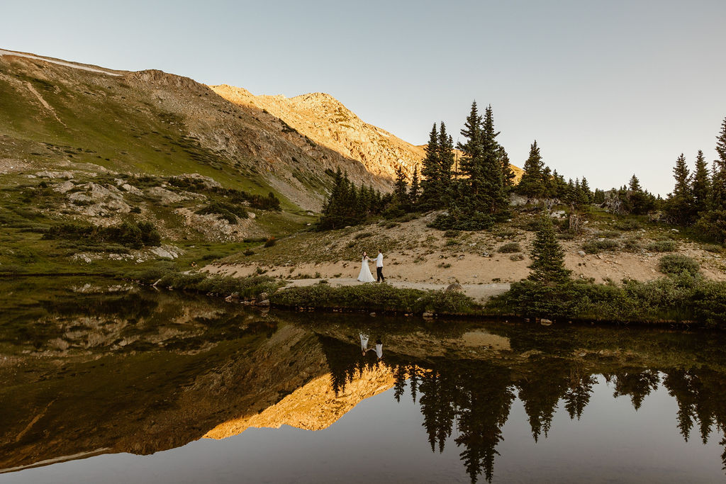a couple is walking holding hands along an alpine lake shore with bright orange alpenglow in the background, and it's all reflecting in the lake