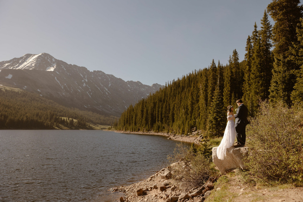 a bride and groom standing on a rock wearing wedding attire next to a lake and a beautiful mountain backdrop in colorado