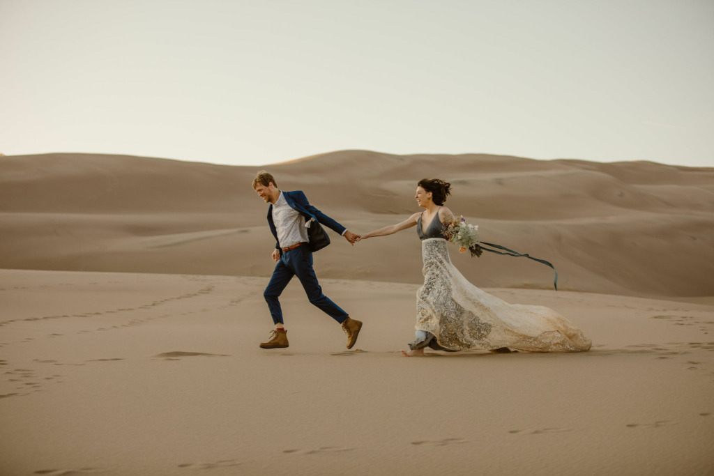 a bride and groom holding hands running through a sand dune