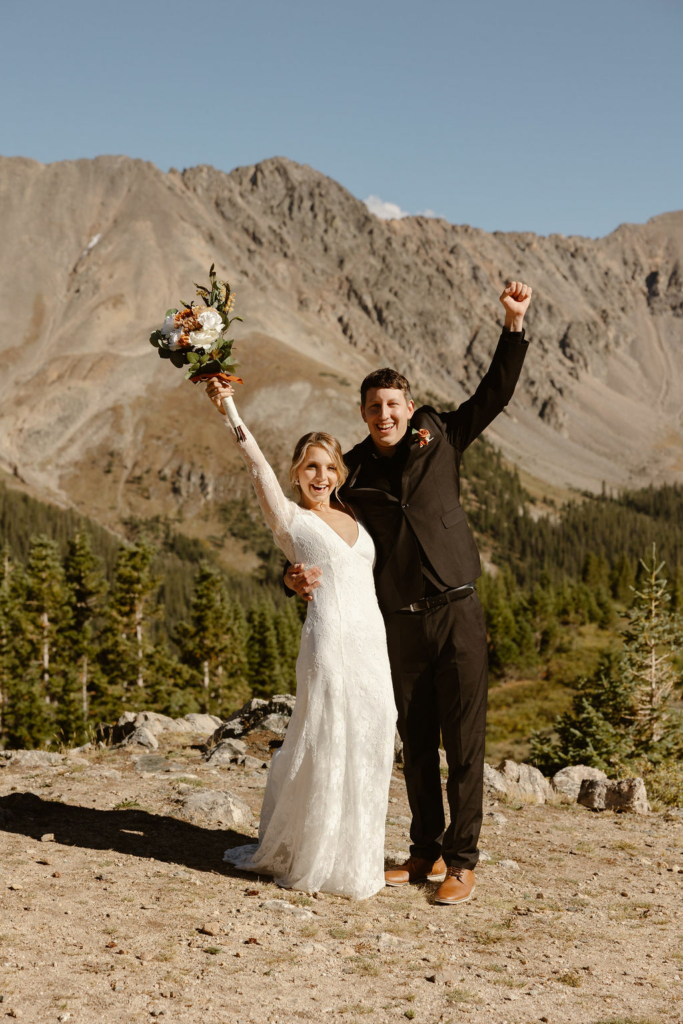 a couple wearing wedding attire celebrating by throwing their hands in the air with a big mountian backdrop during their loveland pass elopement