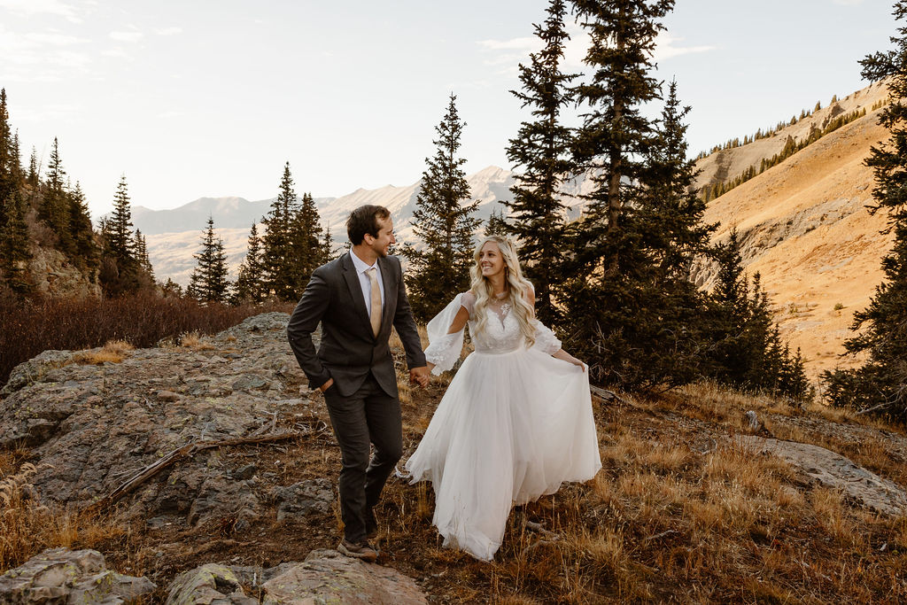 a couple wearing wedding attire holding hands walking towards the camera outdoors during their telluride hiking elopement