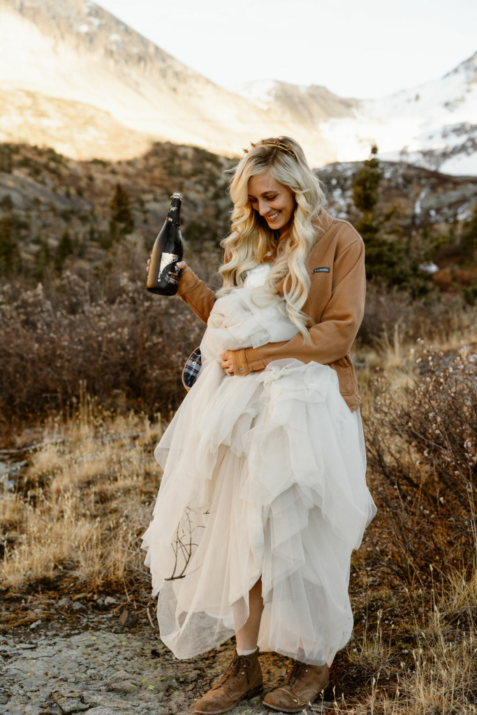 a blonde bride wearing a wedding dress and tan jacket outdoors smiling holding a bottle of champagne