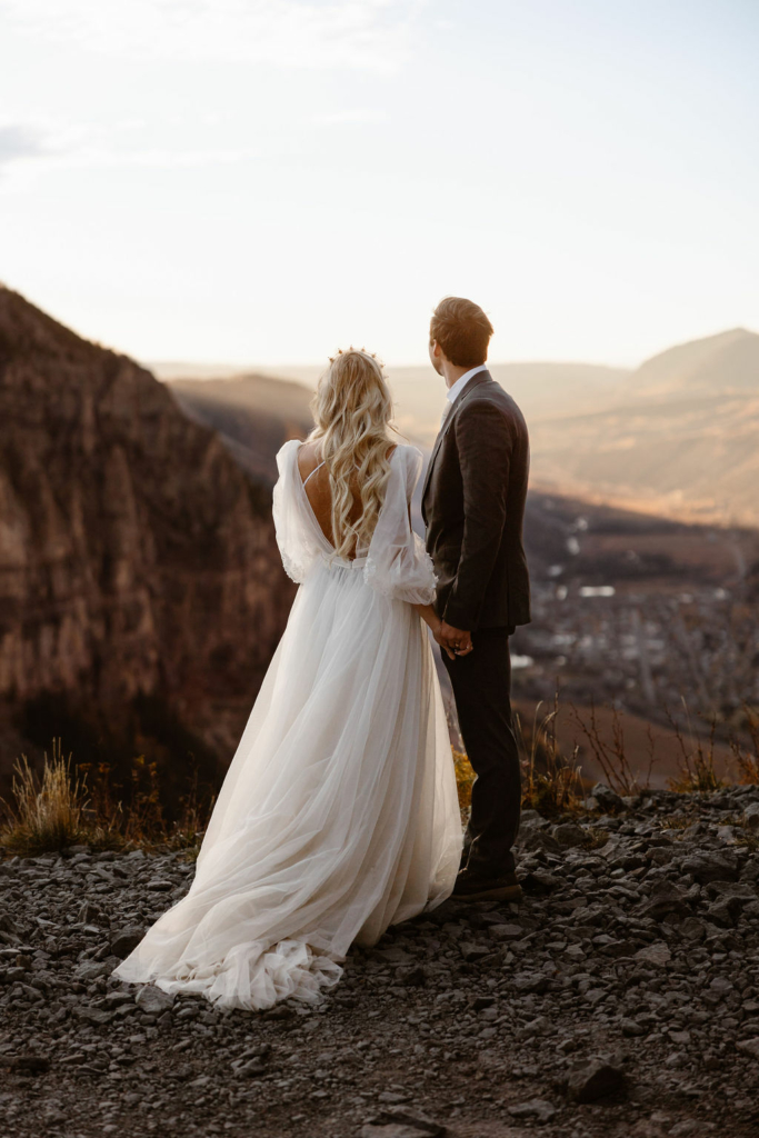 a couple wearing wedding attire sitting at the edge of a cliff overlooking the sunset on telluride, colorado
