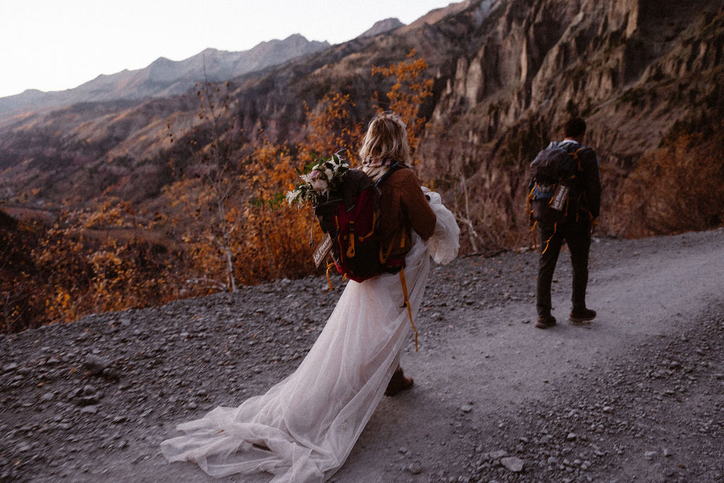 a couple wearing wedding clothes and hiking attire are backpacking down a mountain pass during sunset above telluride, colorado