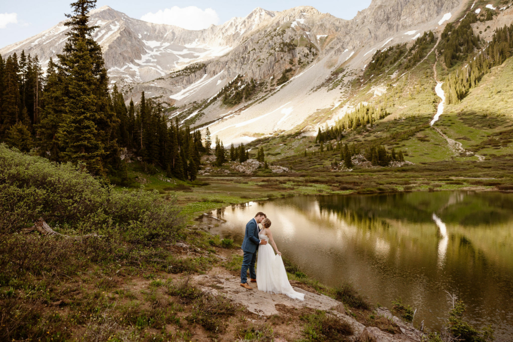 a bride and groom wearing traditional wedding attire kissing next to an alpine lake with beautiful mountain backdrop