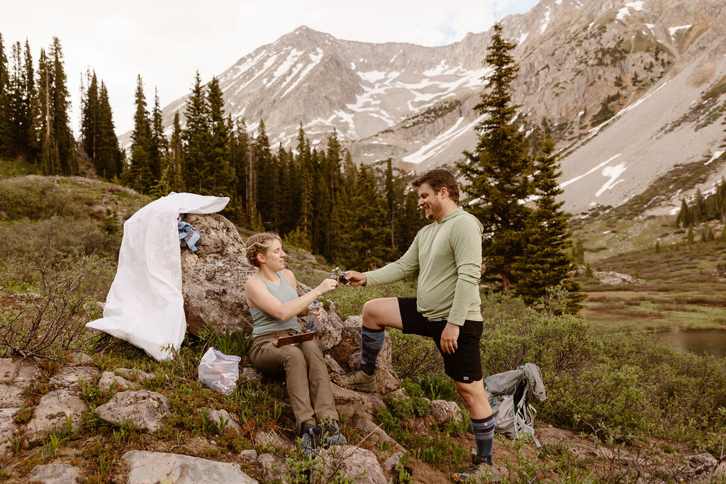 a couple wearing hiking clothes cheersings their shot of whiskey in the mountains during their aspen elopement