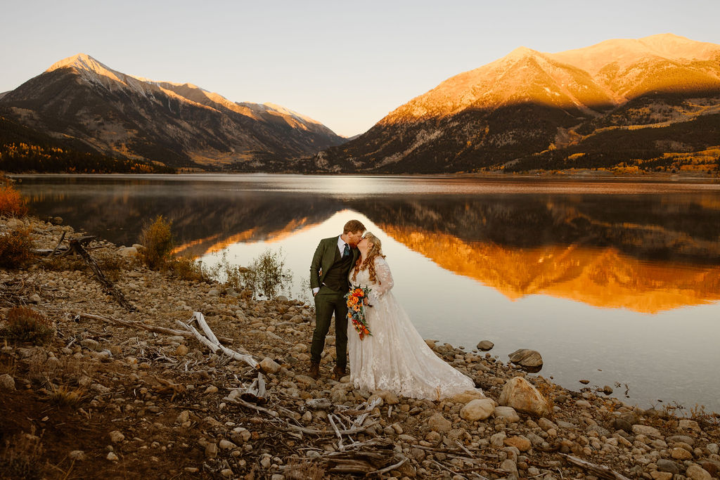 a couple wearing traditional wedding attire kissing in front of a lake with a beautiful mountain backdrop during alpenglow