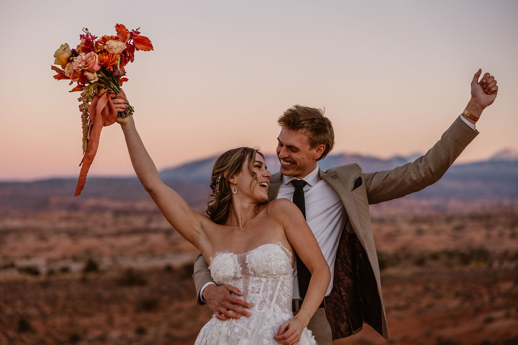 a bride and groom cheering with their arms in the air in a beautiful desert environment