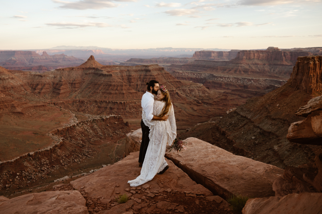 a boho dressed bride and groom standing at the edge of a desert cliff while hugging each other during their canyonlands wedding