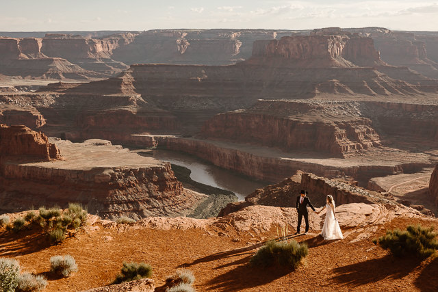 a couple wearing traditional wedding attire holding hands and walking across the cliff side ceremony location during their dead horse point wedding