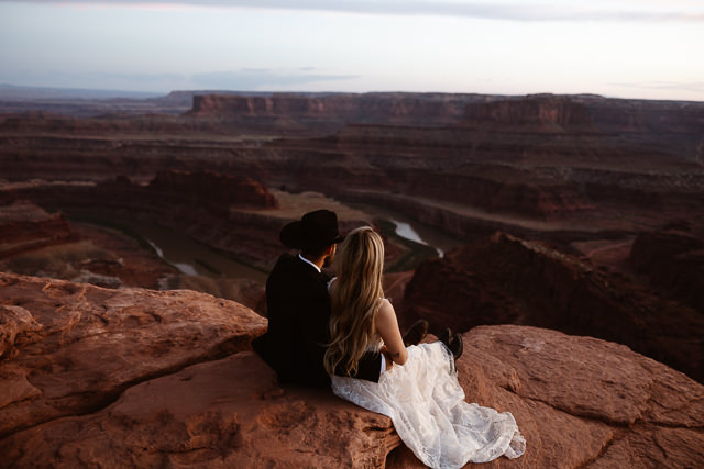 a couple wearing wedding attire sitting on a cliff at dead horse point state park for their wedding