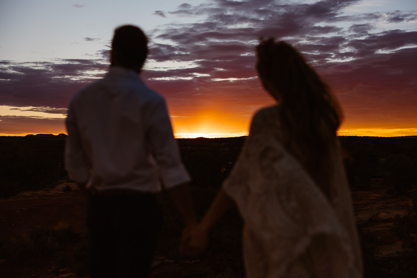 a couple holding hands looking out at a sunset while wearing wedding attire at their dead horse point wedding