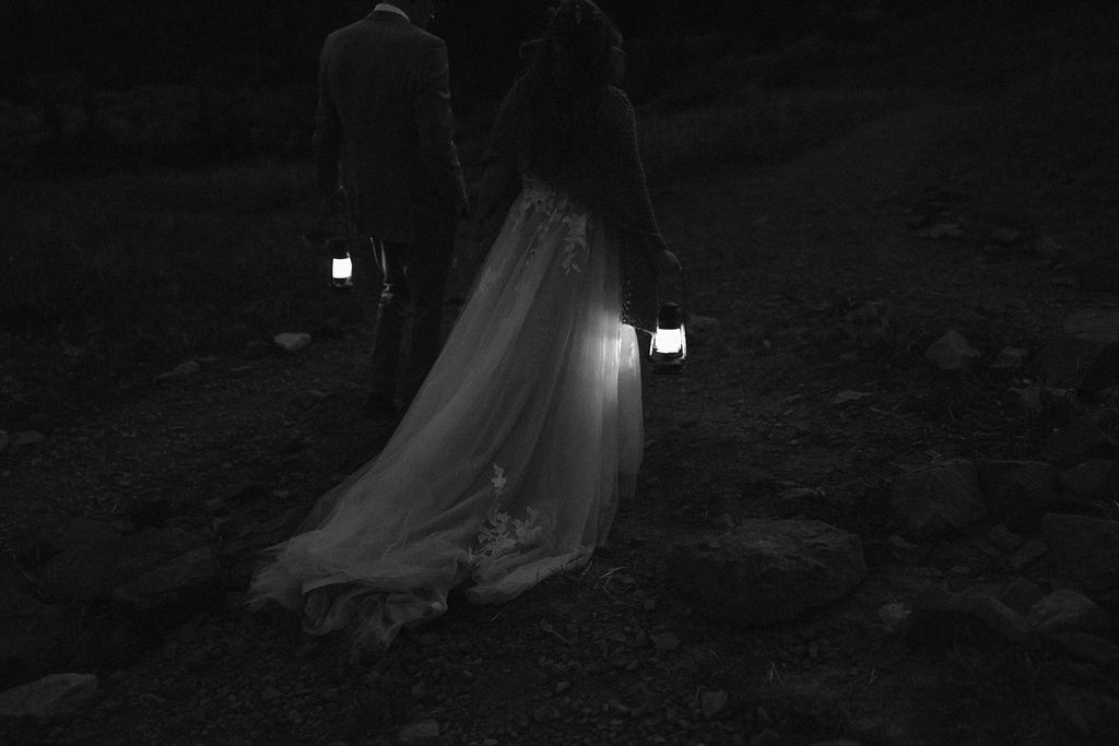 a black and white image of a couple who had an ouray elopement walking into the sunset with two lanterns while wearing wedding attire
