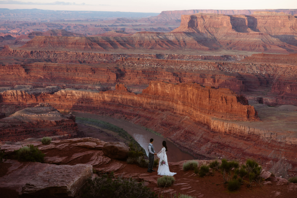 a couple wearing wedding clothes standing at the cliffs edge of dead horse point state park