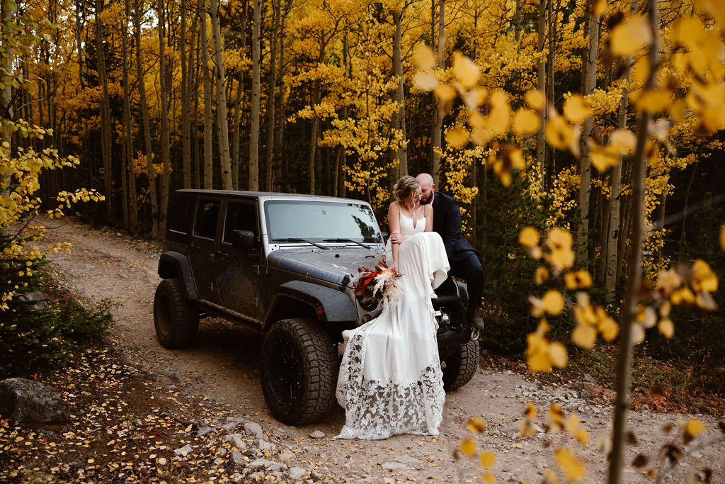 a couple wearing wedding attire sitting on the hood of a Jeep surrounded by golden aspens