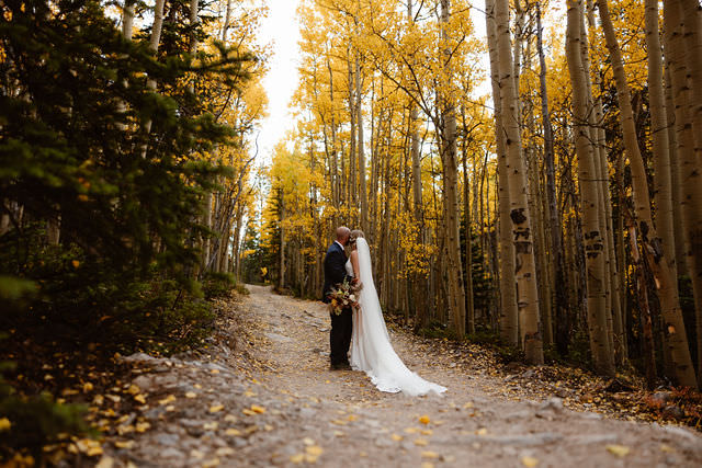 a couple wearing wedding attire standing on a dirt road surrounded by evergreens and golden aspens