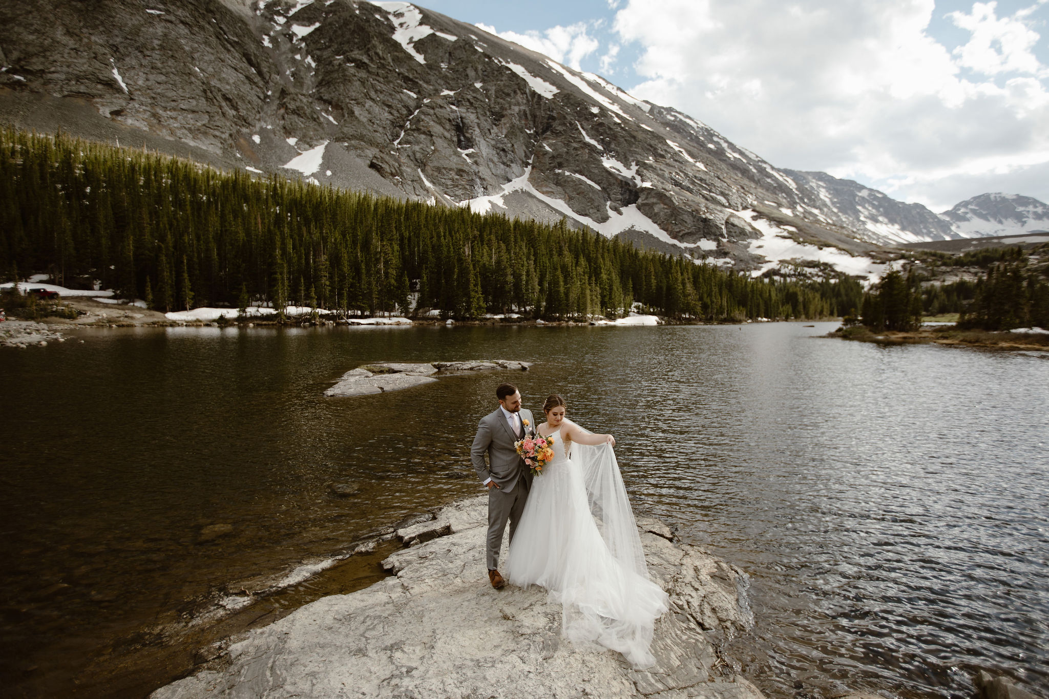 a couple wearing traditional wedding attire standing next to a lake with a big mountain backdrop and blue skys