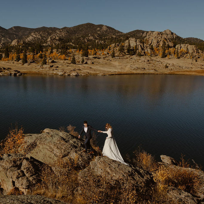 a couple wearing wedding attire and hiking boots walking across a rocky ledge on a lake shore in colorado