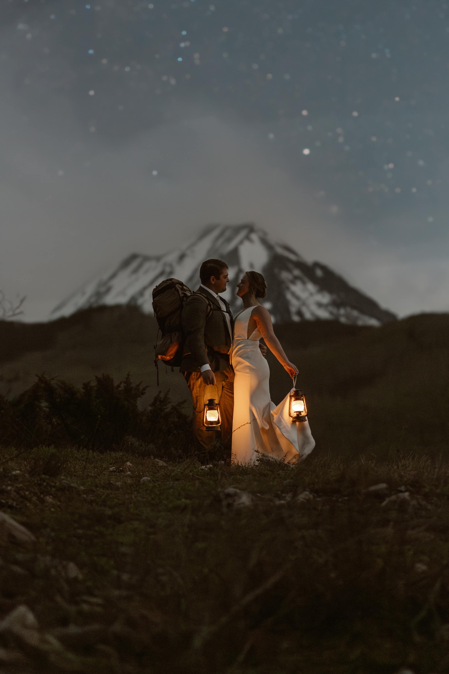 a couple wearing wedding attire standing and kissing while holding lanterns by their sides with a stary night mountain backdrop
