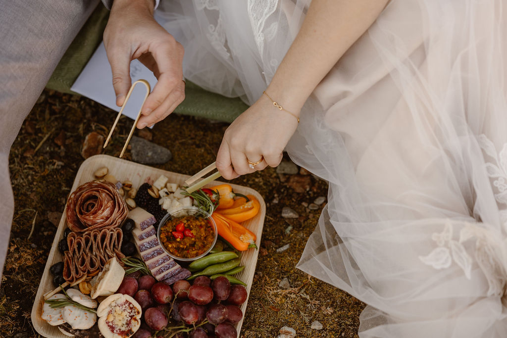 a couple reaching their hands into a charcuterie board while wearing wedding attire during their elopement