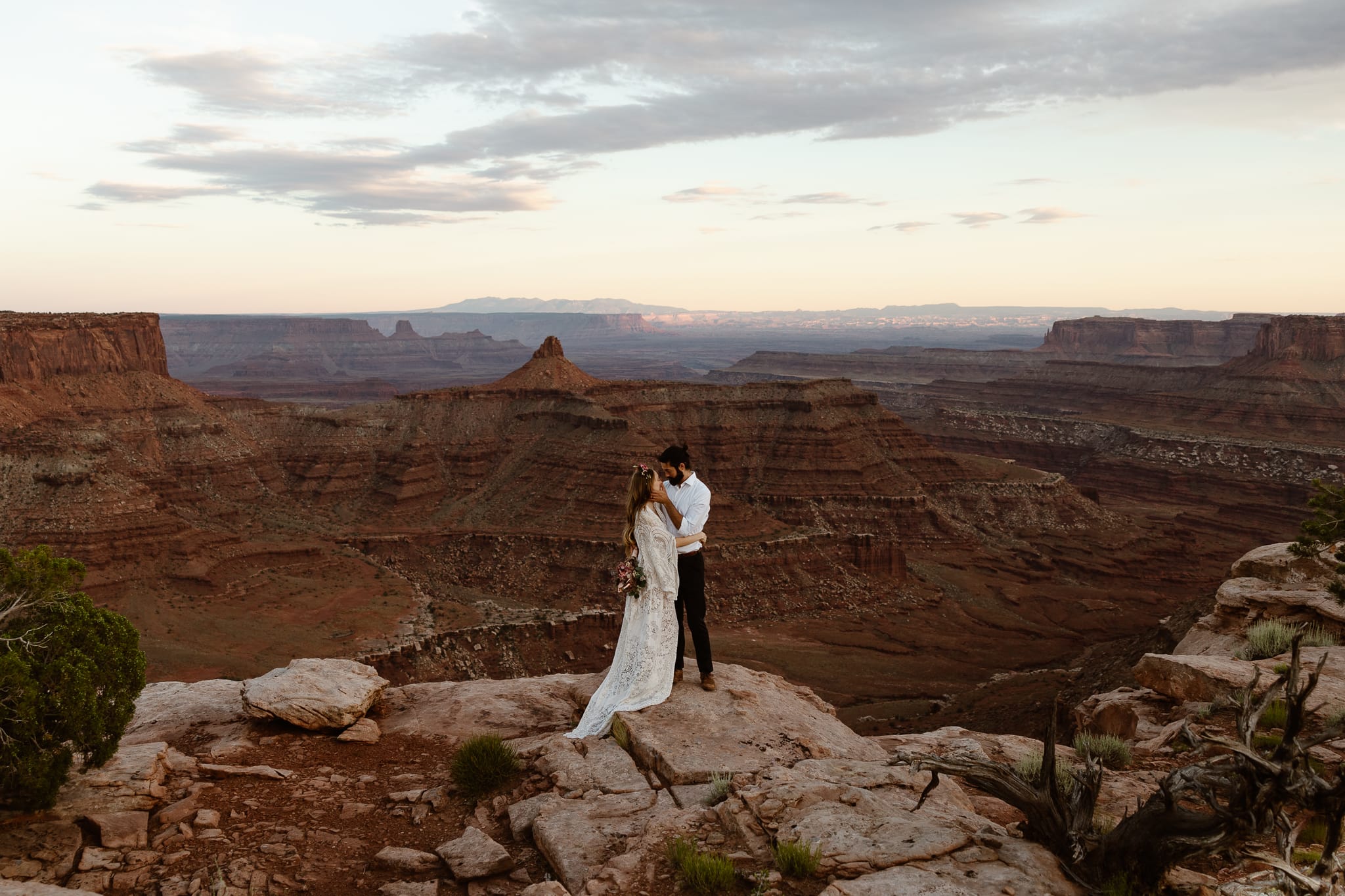 a couple wearing wedding attire standing on the cliffs edge in the desert during sunset