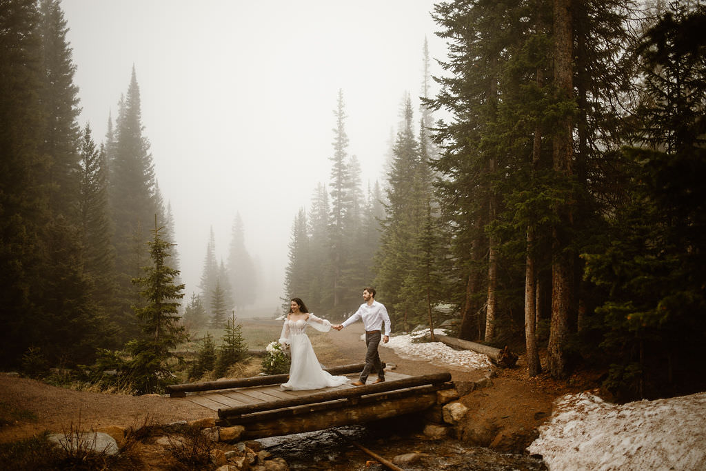 a couple walking over a bridge while holding hands while wearing wedding attire surrounded by fog and rain on their wedding day