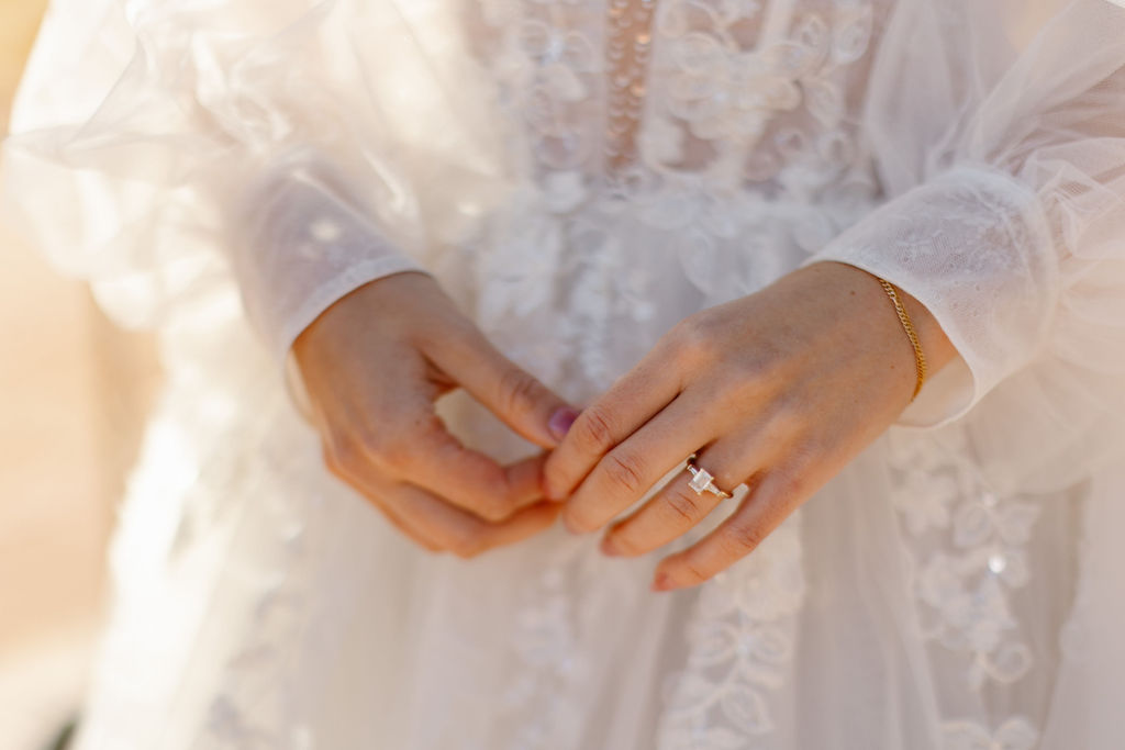 a close up image of a bride's hands, on her left hand is an emerald cut engagement ring and she's wearing long sleeves see through embellished wedding dress